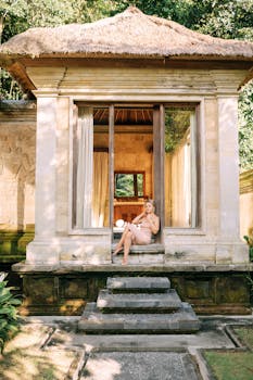 A woman sits elegantly on stone steps of a tranquil stone house with lush greenery.